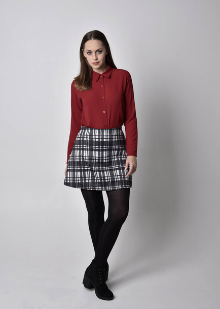full length portrait of a pretty brunette girl wearing a red shirt and plaid skirt with leggings and boots. Standing pose on a  studio background. - Photo, Image