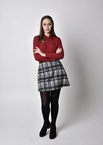 full length portrait of a pretty brunette girl wearing a red shirt and plaid skirt with leggings and boots. Standing pose on a  studio background. - Fotografie, Obrázek