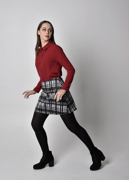 full length portrait of a pretty brunette girl wearing a red shirt and plaid skirt with leggings and boots. Standing pose on a  studio background. - Foto, afbeelding
