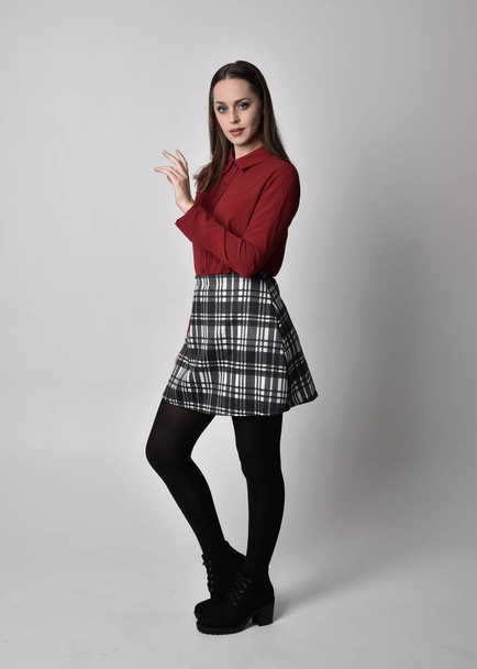 full length portrait of a pretty brunette girl wearing a red shirt and plaid skirt with leggings and boots. Standing pose on a  studio background. - Fotó, kép