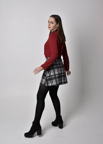 full length portrait of a pretty brunette girl wearing a red shirt and plaid skirt with leggings and boots. Standing pose on a  studio background. - Zdjęcie, obraz