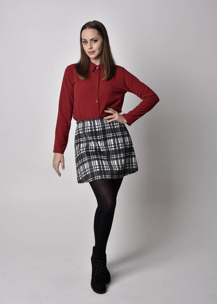 full length portrait of a pretty brunette girl wearing a red shirt and plaid skirt with leggings and boots. Standing pose on a  studio background. - Photo, Image