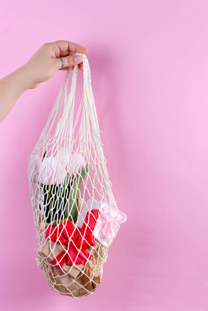 La mano de la hembra sostiene bolsas reutilizables con cajas de regalo de papel, flores frescas de tulipanes de primavera sobre un fondo rosa, espacio para copiar. concepto de bolsa libre de plástico
. - Foto, Imagen