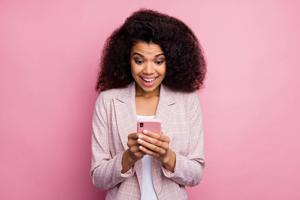 Kuva kaunis innoissaan tumma iho nainen pidä puhelin käsissä lukeminen positiivinen blogikirjoitus kommentit avaa suu kulumista ruudullinen bleiseri eristetty pastelli vaaleanpunainen väri tausta
 - Valokuva, kuva