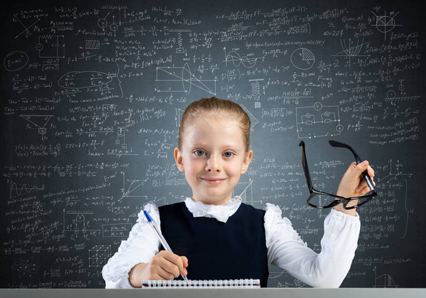 Smiling little girl in schoolwear doing her homework. Time to study concept. Happy schoolgirl with glasses sitting at desk and writing in notebook. Research and education in elementary school. - Φωτογραφία, εικόνα
