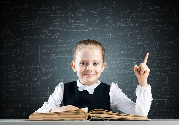 Smiling little girl sitting at desk with open book. Research and education in school. Happy schoolgirl in school wear finger pointing up. Clever girl reading book on chalkboard background. - Photo, Image