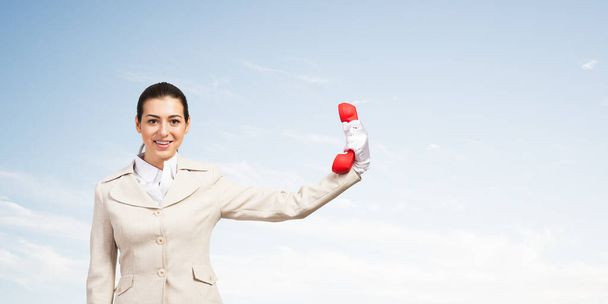 Smiling woman holds red handset phone on distance. Happy operator in business suit posing with cellphone on blue sky background. Business assistance and consultation. Customer service support concept - 写真・画像