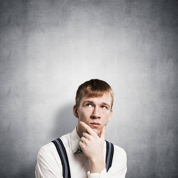 Redhead student looks pensively upwards and tries to remember something. Puzzled boy has serious facial expression. Portrait of guy wears shirt, bow tie and suspenders on background of grey wall. - Fotoğraf, Görsel