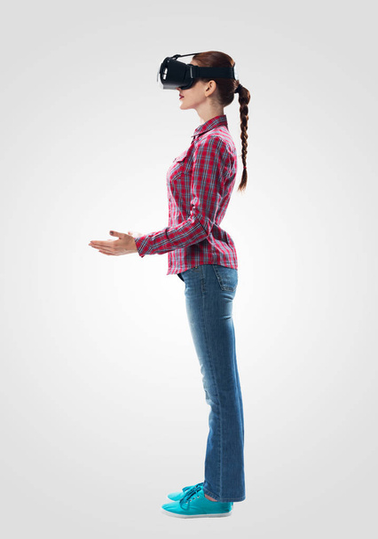 Young woman wearing VR headset and gesturing in air. Woman keep your hands in front of you as something holding. Interacting with digital interface. Studio photo by girl against gray background - Fotografie, Obrázek