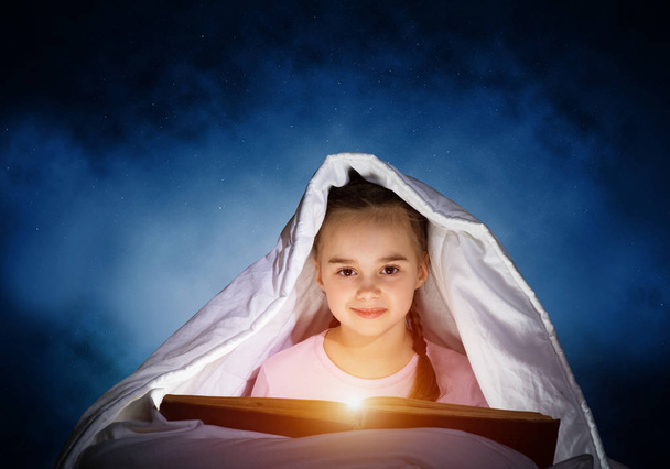 Happy little girl reading interesting book in bed after bedtime. Beautiful child with flashlight hiding under blanket. Smiling girl in pajamas on deep blue sky background. Child reading fairy tales. - Valokuva, kuva