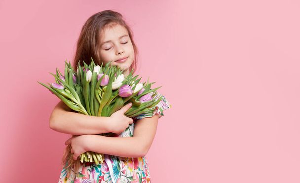 Cute smiling child girl holding bouquet of spring flowers tulips isolated on pink background. Little toddler girl gives a bouquet to mom. Copy space for text.  - Foto, Imagen