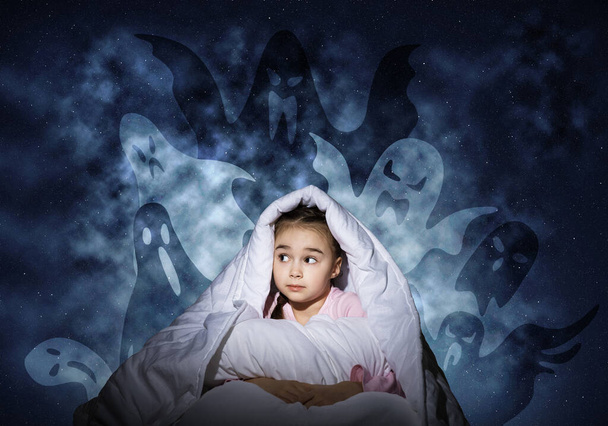Wystraszona dziewczyna chowa się pod kocem. Zaskoczony dzieciak siedzący w łóżku na tle nocnego nieba. Dziewczynka boi się ciemności. Zakryte dziecko nie śpi w nocy. Straszna dziewczyna w piżamie i wymyślonych potworach.. - Zdjęcie, obraz