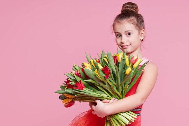 Cute smiling child girl holding bouquet of spring flowers tulips isolated on pink background. Little toddler girl gives a bouquet to mom. Copy space for text.  - Foto, imagen