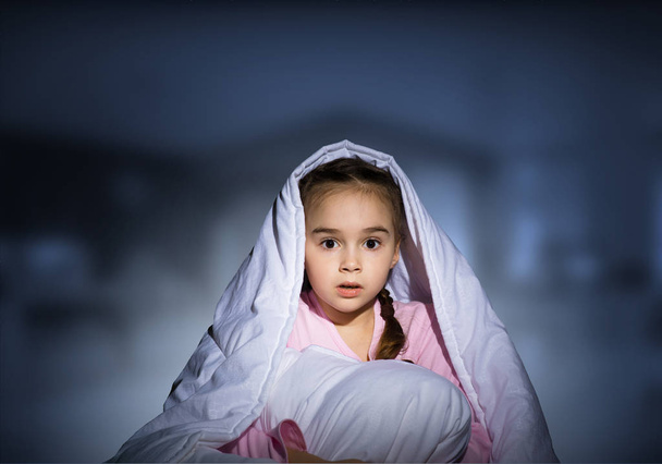 Scared girl with pillow hiding under blanket. Startled kid lying in his bed at home. Little girl afraid of dark. Covered child not sleep at night. Girl in pajamas on deep blue blurred background - Фото, зображення
