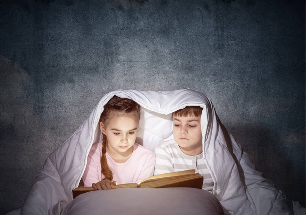 Little girl and boy looking at each other under blanket. Children reading magic stories in bed before going to sleep. Young sister and brother in pajamas together on background of grey wall. - Photo, Image