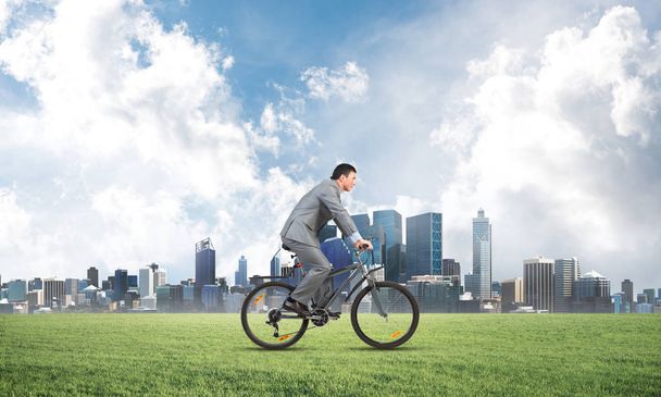 Side view of businessman riding bicycle on green field. Man in business suit on bike outdoor. Cyclist on background of countryside landscape with blue sky and city on horizon. Eco-friendly transport. - Foto, afbeelding