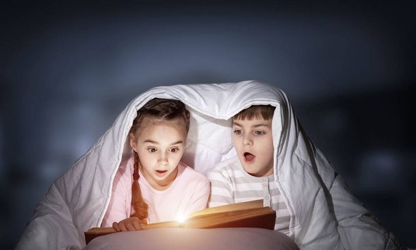 Children reading scary stories in bed before going to sleep. Little girl and boy in pajamas hiding under blanket together. Covered kids with open book not sleep at night. Fear of the dark concept - Φωτογραφία, εικόνα