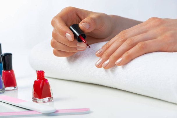 Woman giving herself elegant manicure at home. Closeup of beautiful female hand applying red nail polish at white table. Professional nail care and beautician procedure. Stylish nail art concept - Φωτογραφία, εικόνα