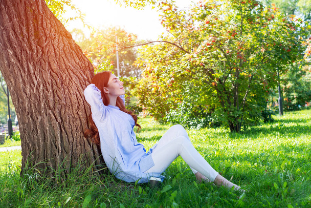 Young woman sitting under tree in summer park on sunny day. Beautiful redhead girl relaxing with closed eyes. Happy lady sitting on green grass leaning against tree. Healthy lifestyle and relaxation. - Photo, image