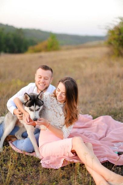 Young european man and girl sitting on pink plaid outside and playing with husky. - 写真・画像