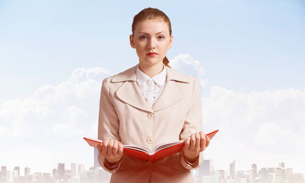Attractive woman holding open notebook on background of cityscape and blue sky. Elegant young woman in white business suit with open book in hands. Internship and practice in company. - Fotoğraf, Görsel