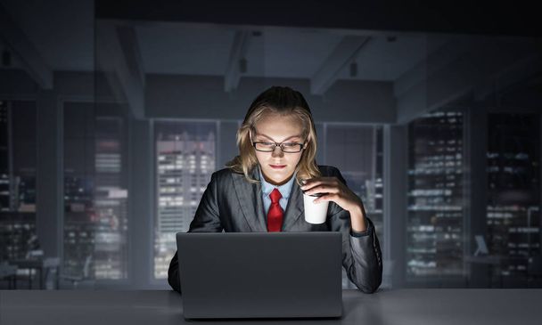 Businesswoman sitting at desk and looking at laptop. Secretary in business suit and tie with cup of coffee. Working in office late at night. Overwork and deadline concept with corporate employee - Foto, Imagen