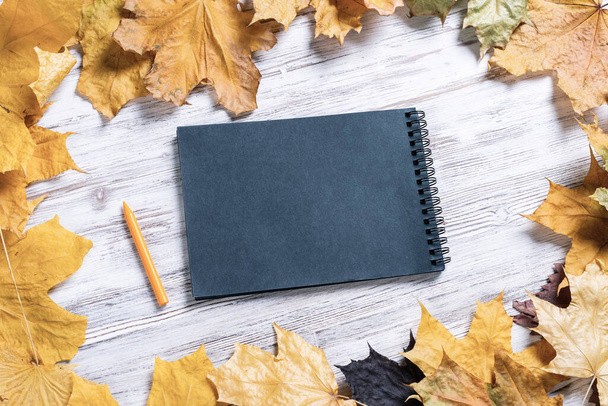 Spiral black notepad and pen lies on vintage wooden desk with bright autumn foliage. Business and education. Flat lay with autumn leaves on white wooden surface. Blank notepaper with copy space. - Φωτογραφία, εικόνα