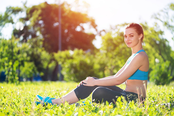 Beautiful smiling girl in activewear relax in park at sunrise. Portrait of young charming woman sitting on green grass. Training and meditation outdoor. Morning exercises and healthy lifestyle. - Zdjęcie, obraz