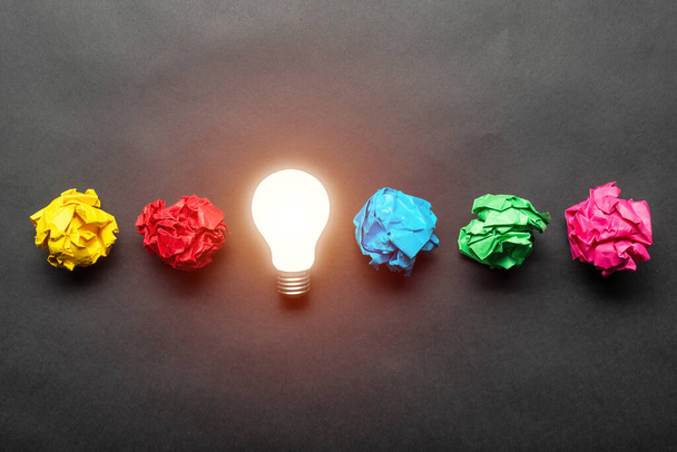 Lightbulb and crumpled colorful paper balls on black background. Successful solution of problem. Idea generation and brainstorming. Genius idea among failing ideas metaphor. Business motivation - Photo, Image