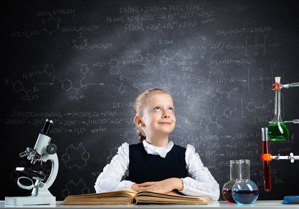 Schoolgirl sitting at desk with open book. Happy girl scientist studying in classroom with chalkboard. Chemical laboratory with glass flasks and test tubes. Clever girl in schoolwear reading book on - Foto, Imagen