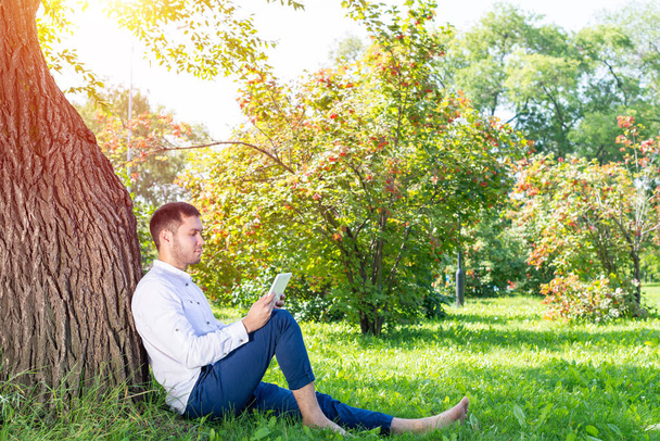 Young man using tablet computer under tree in park on sunny day. Handsome businessman looking at tablet screen. Calm man sitting on green grass leaning against tree. Mobile communication concept. - Foto, Imagem