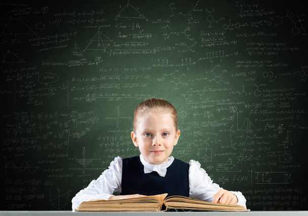 Smiling little girl sitting at desk with open book. Research and education in school. Happy schoolgirl studying in library. Clever girl in schoolwear reading book on chalkboard background. - Foto, immagini