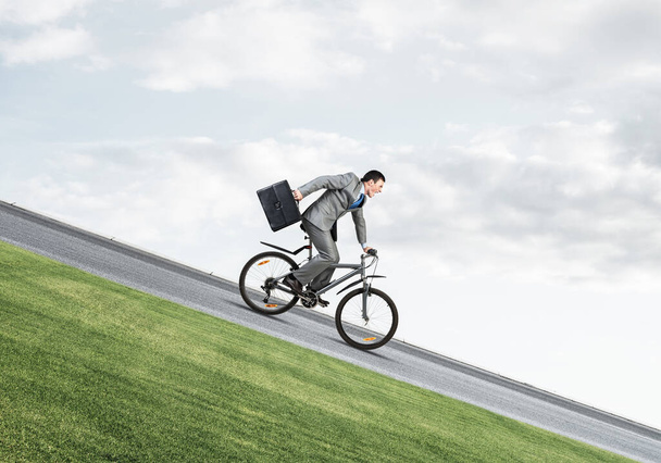 Young man on bicycle fast riding downhill at sunny day. Businessman on bike hurry to work. Corporate employee wearing business suit with suitcase on cycle outdoor. Male cyclist on mountain road - Фото, изображение