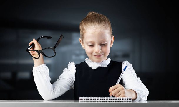 Smiling little girl in schoolwear doing her homework. Time to study concept. Happy schoolgirl with glasses sitting at desk and writing in notebook. Research and education in elementary school. - Photo, Image