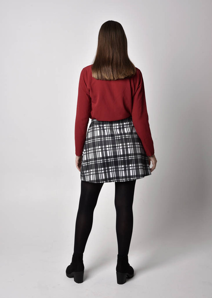 full length portrait of a pretty brunette girl wearing a red shirt and plaid skirt with leggings and boots. Standing pose facing away from the camera on a  studio background. - Zdjęcie, obraz