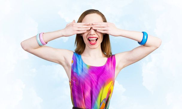 Redhead beautiful girl closing her eyes with hands. Charming lady wears bright dress and bracelets standing against blue sky background. Portrait of happy young woman waiting for pleasant surprise. - Foto, Imagen