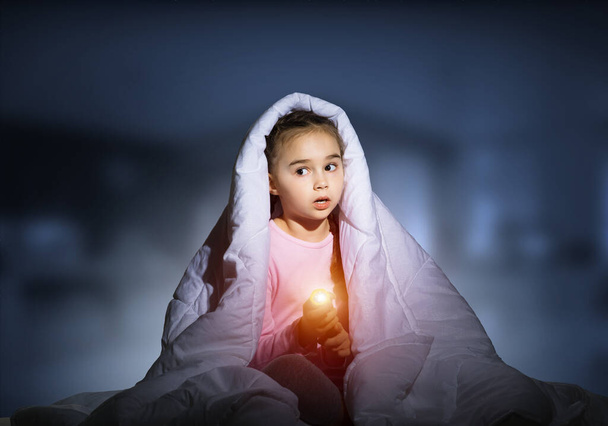 Frightened girl with flashlight hiding under blanket. Scared kid lying in his bed at home. Fear of the dark. Beautiful little girl not sleep at night. Child in pajamas on blue blurred background. - Фото, зображення