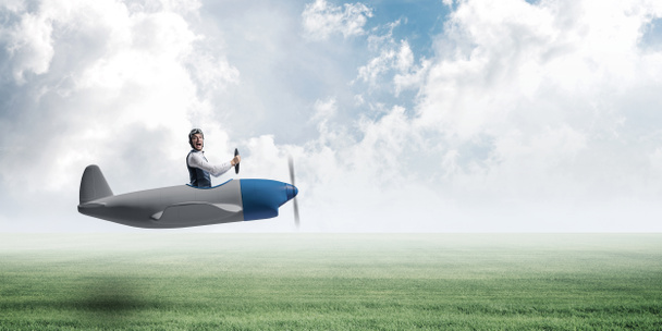 Happy aviator driving propeller plane. Emotional pilot in leather helmet looking in camera. Man in airplane flying low above green meadow. Nature panorama with green grass and cloudy sky in summer day - Photo, Image