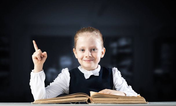 Smiling little girl sitting at desk with open book. Research and education in school. Happy schoolgirl in school wear finger pointing up. Clever girl reading book on dark blurred background. - Foto, Imagem