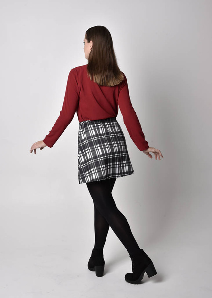 full length portrait of a pretty brunette girl wearing a red shirt and plaid skirt with leggings and boots. Standing pose facing away from the camera on a  studio background. - Photo, Image