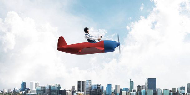 Businessman in aviator hat and goggles driving propeller plane above downtown with high modern buildings. Successful leader flying in small airplane. Blue sky landscape with clouds on background - Photo, Image