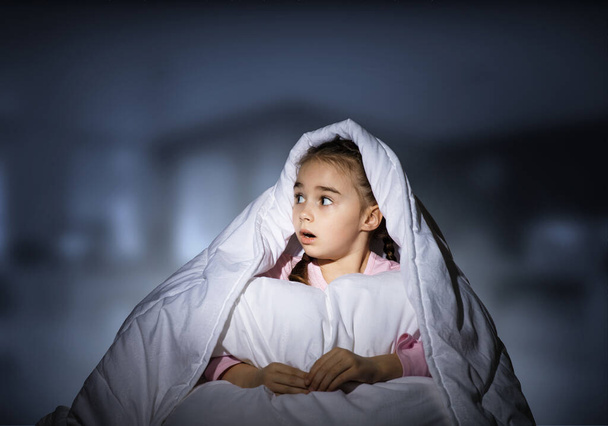 Scared girl with pillow hiding under blanket. Startled kid lying in his bed at home. Little girl afraid of dark. Covered child not sleep at night. Girl in pajamas on blue blurred background. - Foto, Imagem