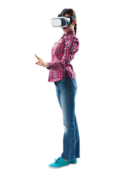 Young woman wearing VR goggles and gaming in interactive game. Lady in checkered shirt and jeans gesturing in air. Studio photo by girl against gray background. Digital entertainment in cyberspace - Photo, image