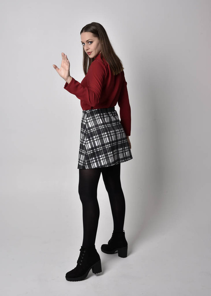 full length portrait of a pretty brunette girl wearing a red shirt and plaid skirt with leggings and boots. Standing pose facing away from the camera on a  studio background. - Photo, Image