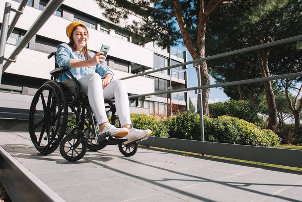 Κορίτσι με ειδικές ανάγκες σε αναπηρικό καροτσάκι γελώντας ενώ χρησιμοποιεί το κινητό της τηλέφωνο σε μια ηλιόλουστη μέρα. - Φωτογραφία, εικόνα