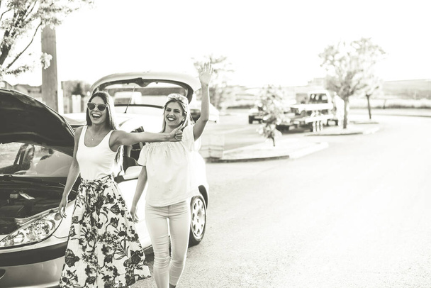 Δύο νεαρές γυναίκες προσπαθούν να σταματήσουν ένα αυτοκίνητο, καθ 'οδόν. Το αμάξι τους είναι χαλασμένο και χρειάζονται βοήθεια. Αλλά είναι χαρούμενα και διασκεδάζουν. Ασπρόμαυρη φωτογραφία. - Φωτογραφία, εικόνα