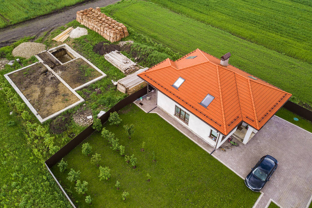 Aerial top view of a private house with attic windows on roof, paved yard with green grass lawn and building site with concrete foundation floor and stacks of yellow bricks for construction.. - Photo, Image