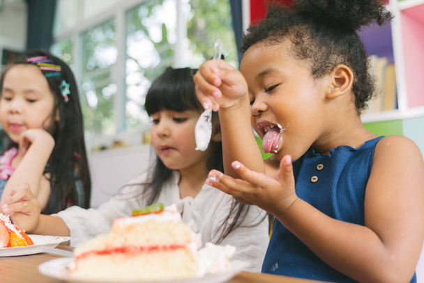 cute little child girl with diversity friends eating cake together. kids eat dessert . - Zdjęcie, obraz