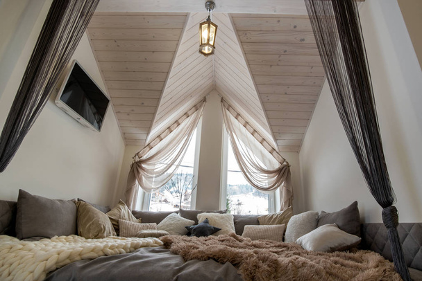 大きな柔らかい休憩所とモダンな家の広々とした廊下のインテリア。木製のロフト天井の上に多くの枕やライトウィンドウと現代的な広いソファ. - 写真・画像
