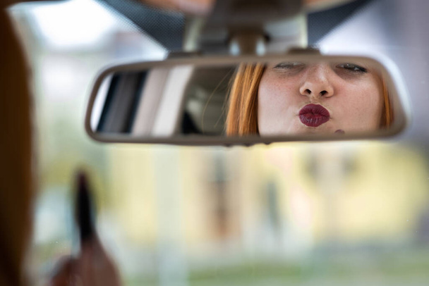 Close-up van een jonge roodharige vrouw bestuurder corrigeren van haar make-up met donkerrode lippenstift kijken in auto achteruitkijkspiegel achter stuurwiel van een voertuig. - Foto, afbeelding